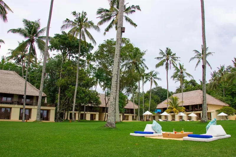Des bungalows aux toits de chaume s'alignent sur une pelouse verdoyante bordée de palmiers.