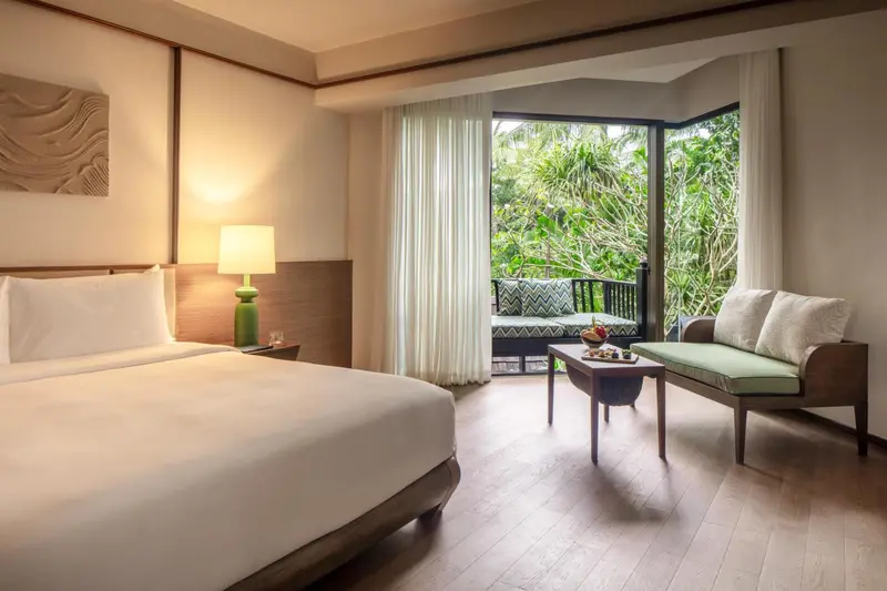 Une chambre lumineuse avec un grand lit et un coin salon donnant sur une végétation luxuriante.