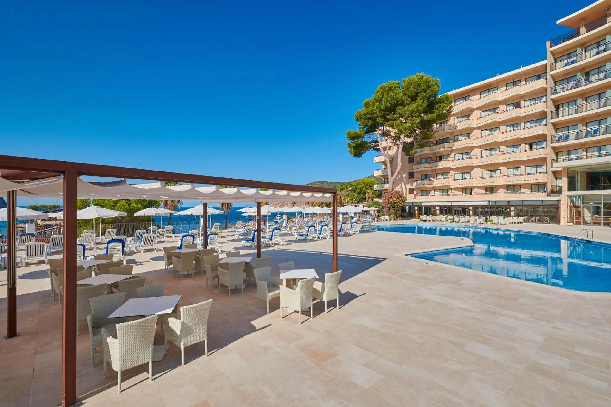 Terrasse ombragée avec tables et chaises donnant sur une piscine et la mer.