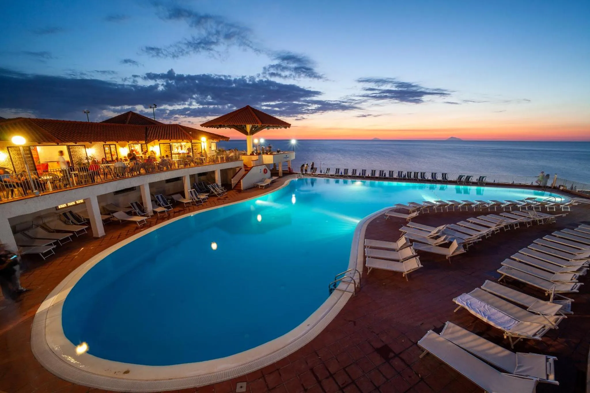 Piscine illuminée avec transats et vue sur la mer au coucher du soleil.