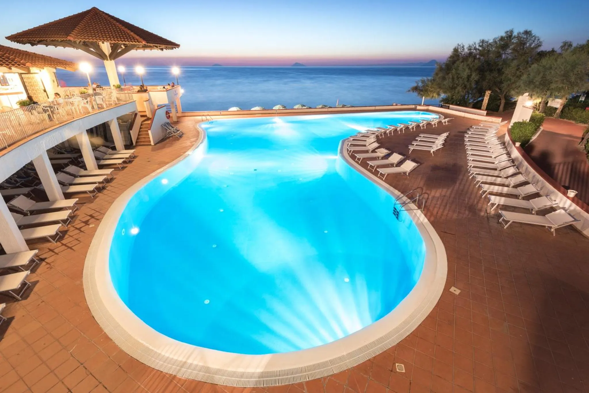 Piscine illuminée avec transats et vue sur la mer au coucher du soleil.