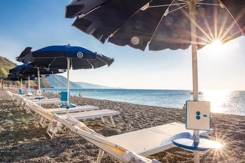 Transats blancs alignés sur une plage de galets sous des parasols noirs.