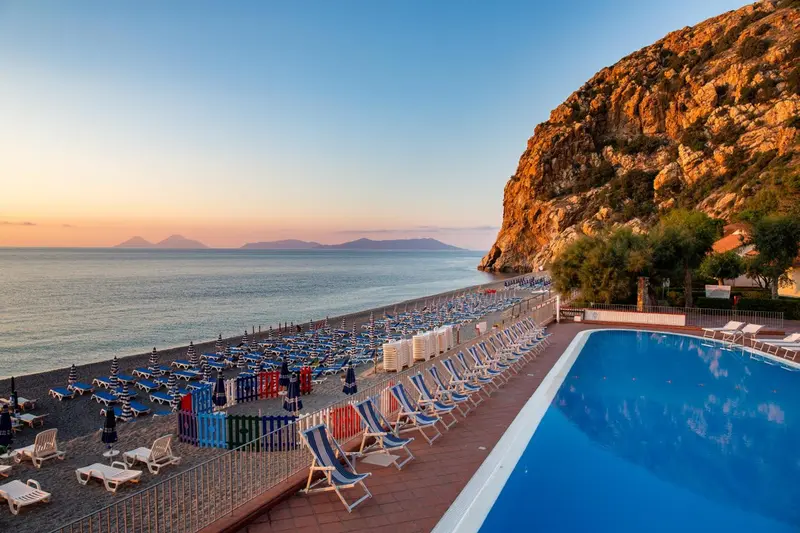 Une piscine turquoise bordée de transats, avec une plage et la mer en arrière-plan.