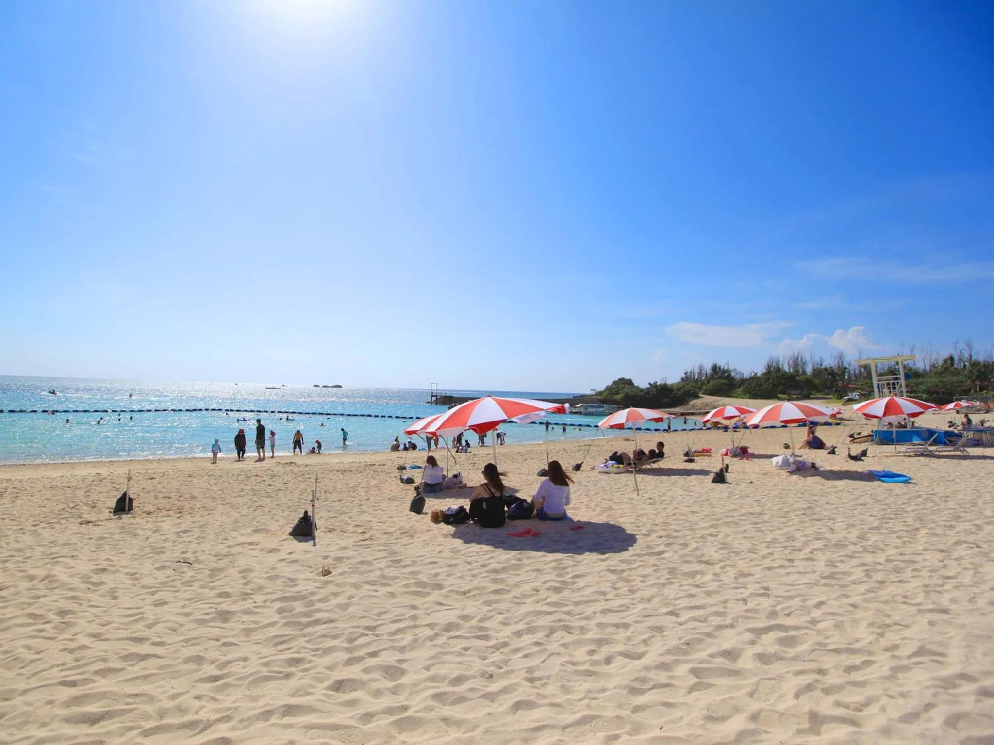 Plage de sable fin avec parasols rouges et blancs et personnes se relaxant au soleil.