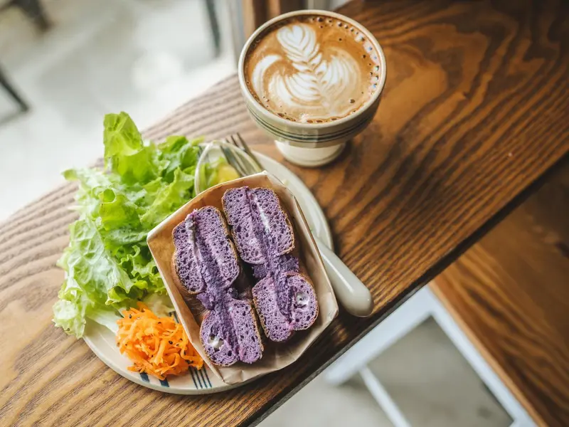Une table en bois présente une tasse de café et une assiette avec des sandwichs violets, de la salade et des carottes râpées.