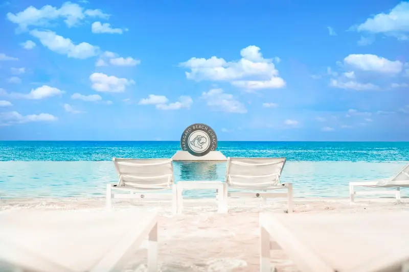 Deux transats blancs invitent à la détente sur une plage de sable fin face à une mer turquoise.