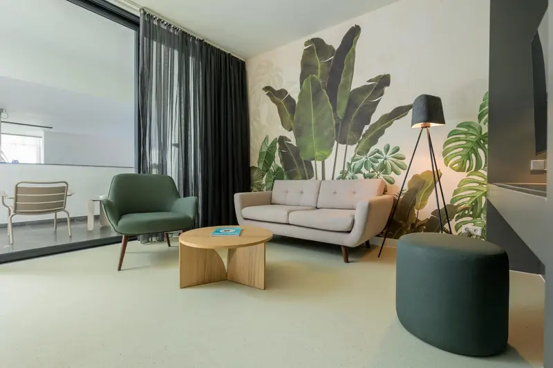 Un salon moderne avec canapés verts et gris, une table basse en bois et un grand motif de feuilles tropicales sur le mur.