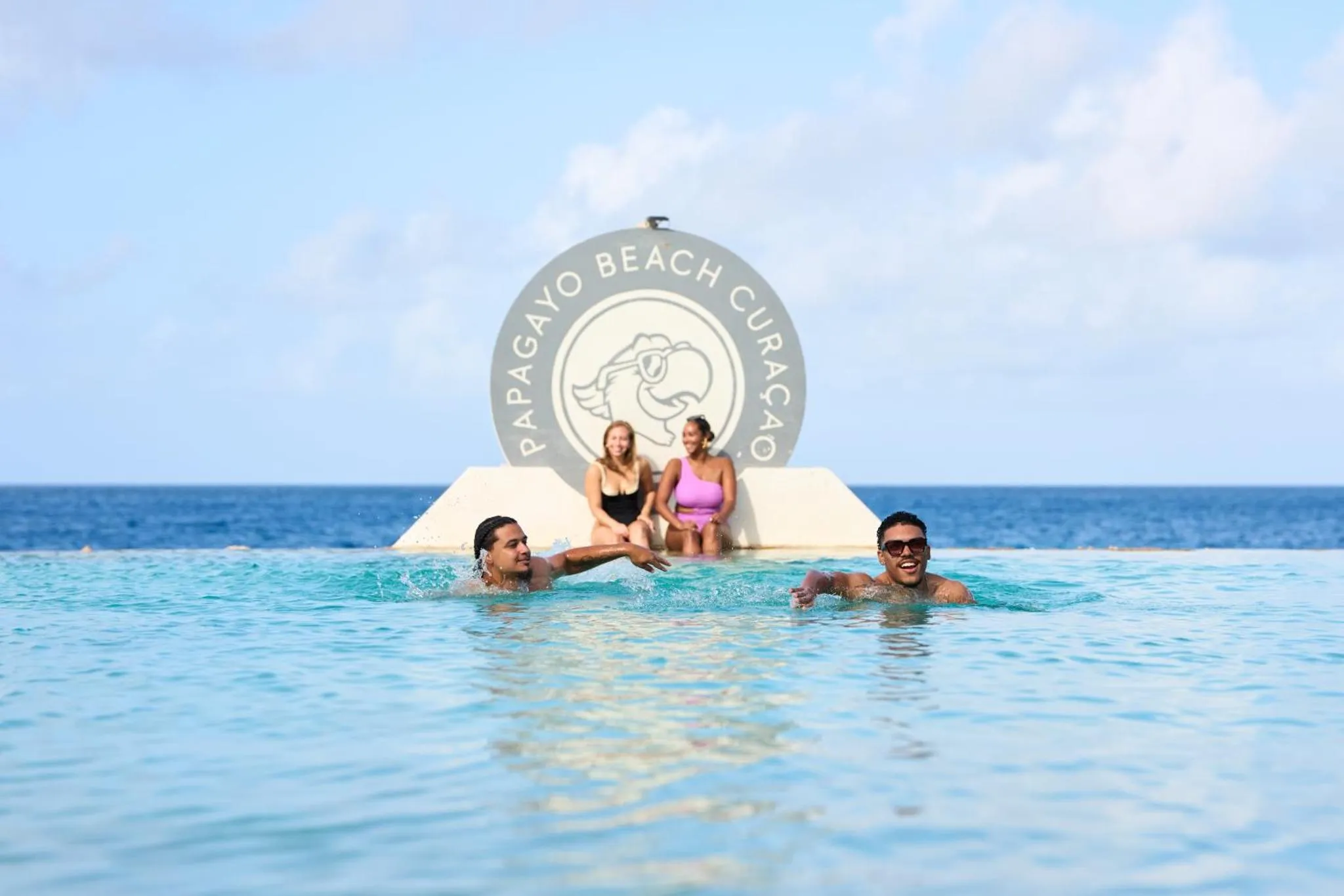 Des personnes sourient et s'amusent dans une piscine turquoise face à la mer.
