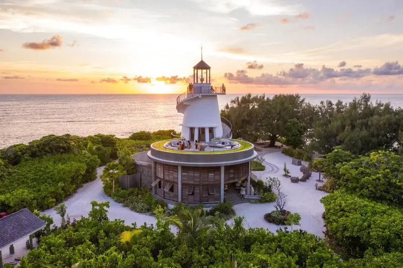 Un phare blanc s'élève au milieu d'une végétation luxuriante face à la mer au coucher du soleil.