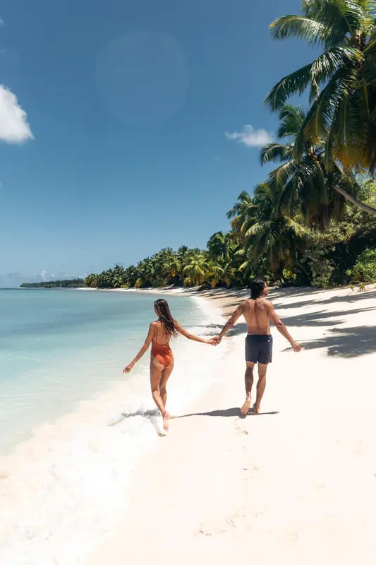 Un couple se promène main dans la main sur une plage de sable blanc bordée de palmiers.