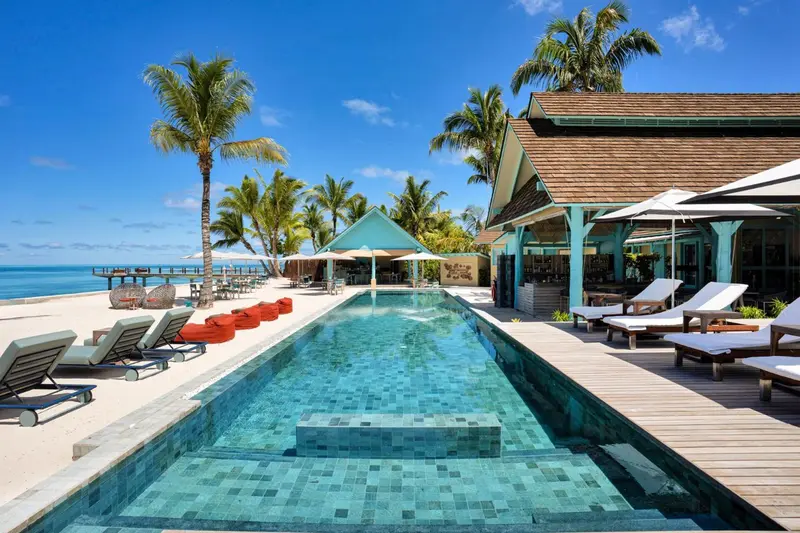 Une piscine turquoise bordée de transats et de palmiers, avec un bâtiment coloré en arrière-plan.