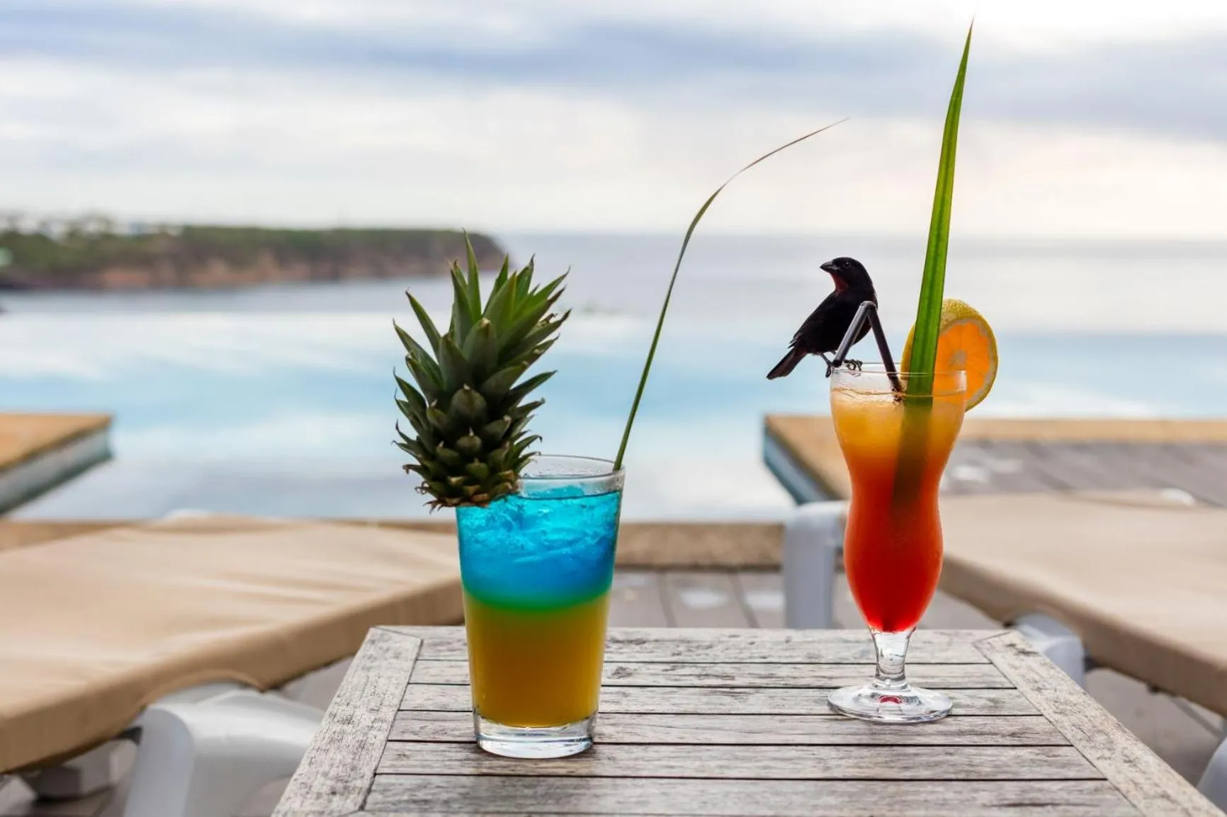 Deux cocktails colorés décorés de fruits exotiques sont présentés sur une table en bois au bord d'une piscine.