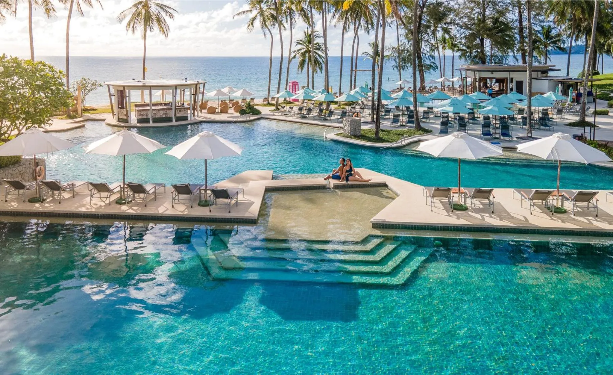 Une piscine turquoise bordée de transats et de parasols blancs, avec une vue sur la mer.