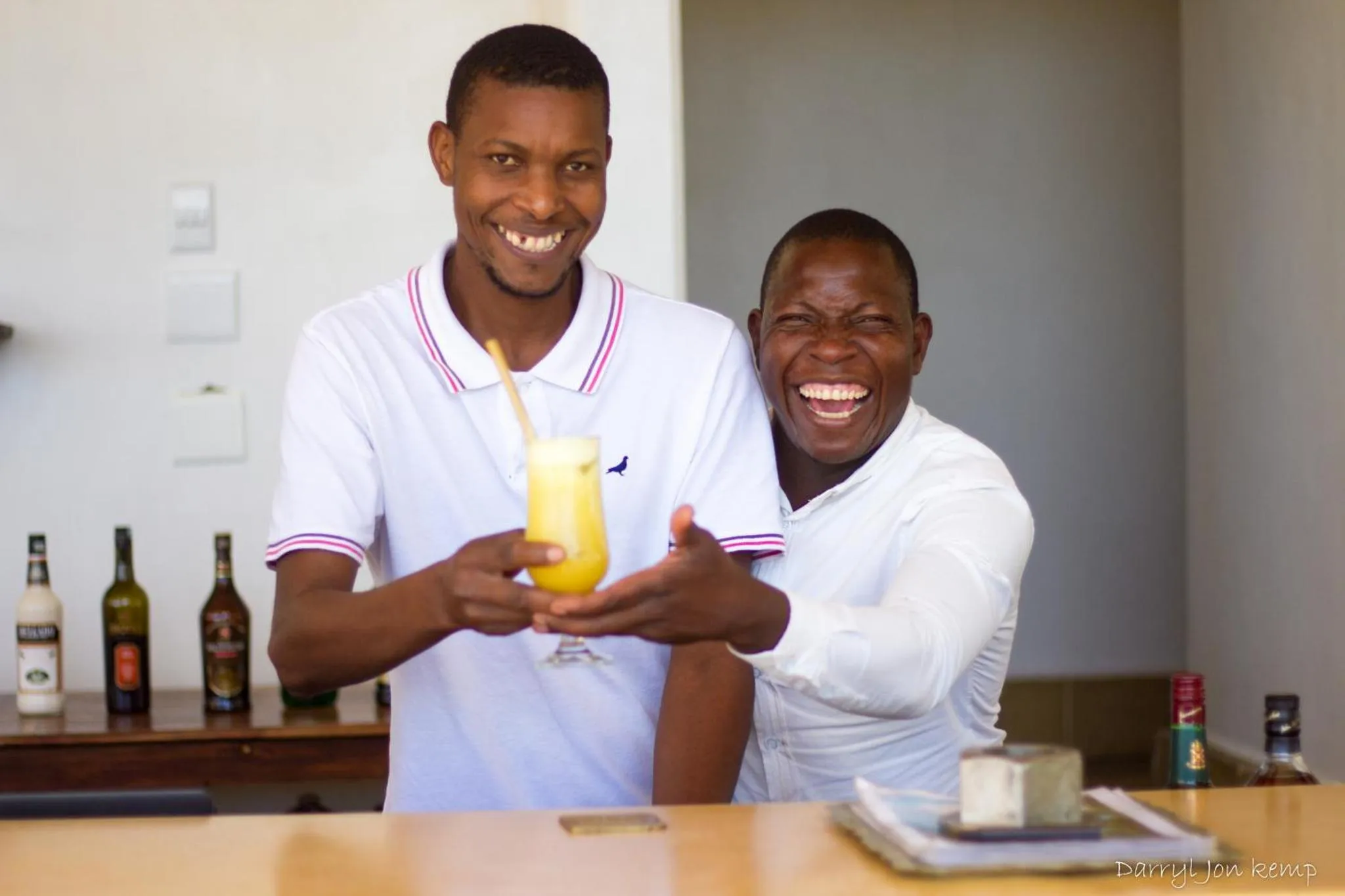 Deux hommes souriants derrière un comptoir de bar présentent un cocktail.