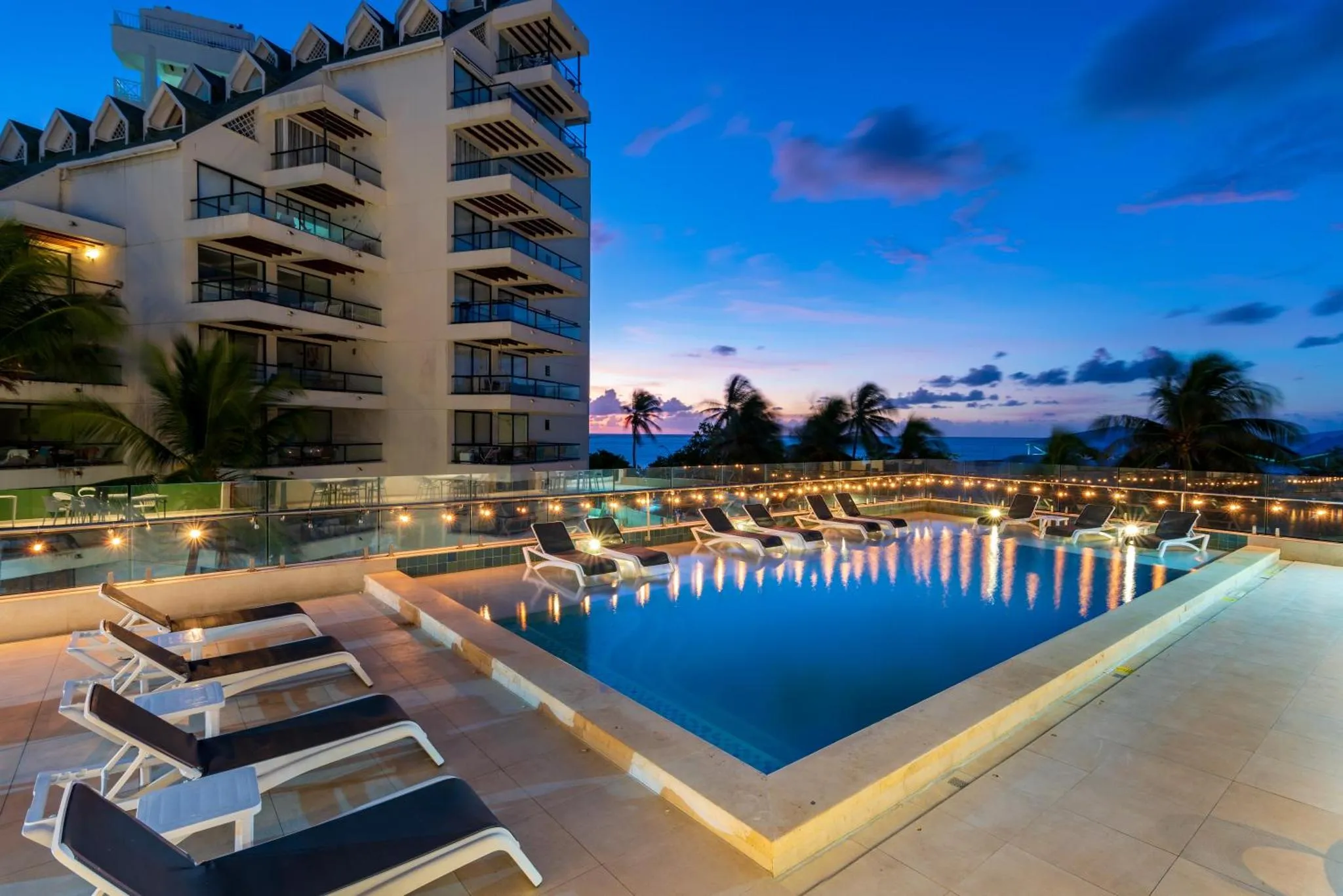 Piscine rectangulaire avec transats et bâtiment en arrière-plan au coucher du soleil.