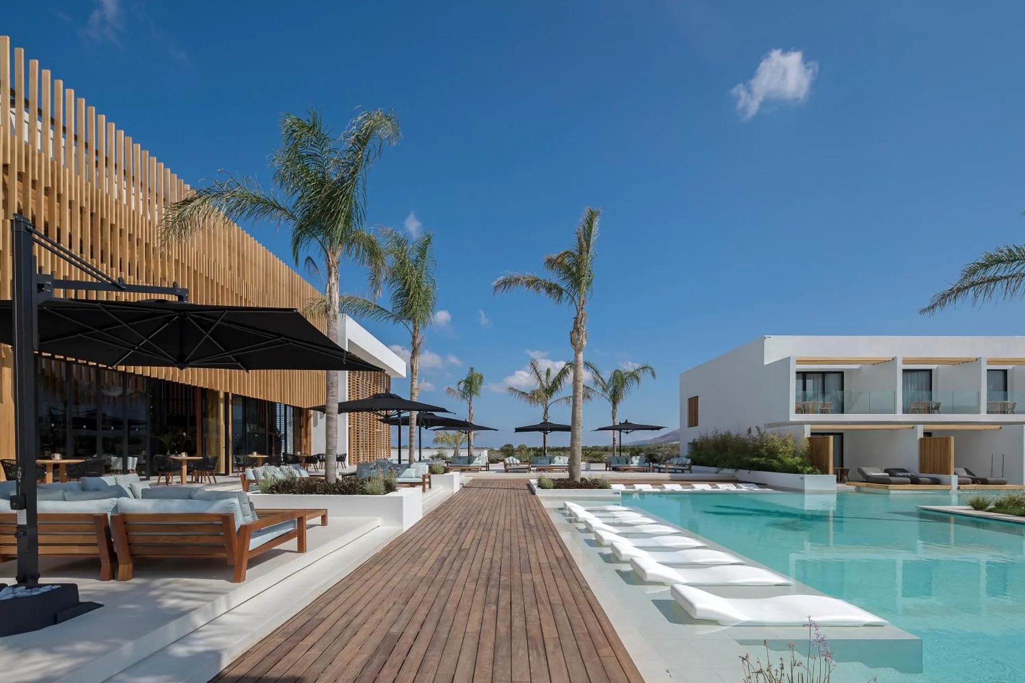 Terrasse en bois avec mobilier de détente et piscine turquoise devant un bâtiment moderne.