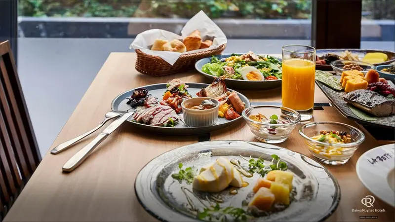 Une table en bois présente un copieux petit-déjeuner avec des viennoiseries, des fruits, des œufs et des salades.