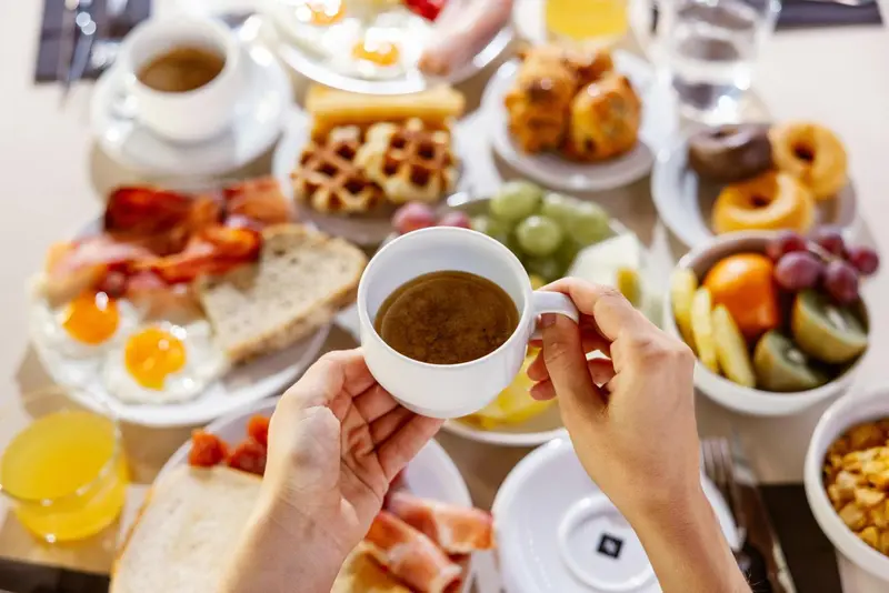 Une main tenant une tasse de café au-dessus d'une table garnie de mets variés.