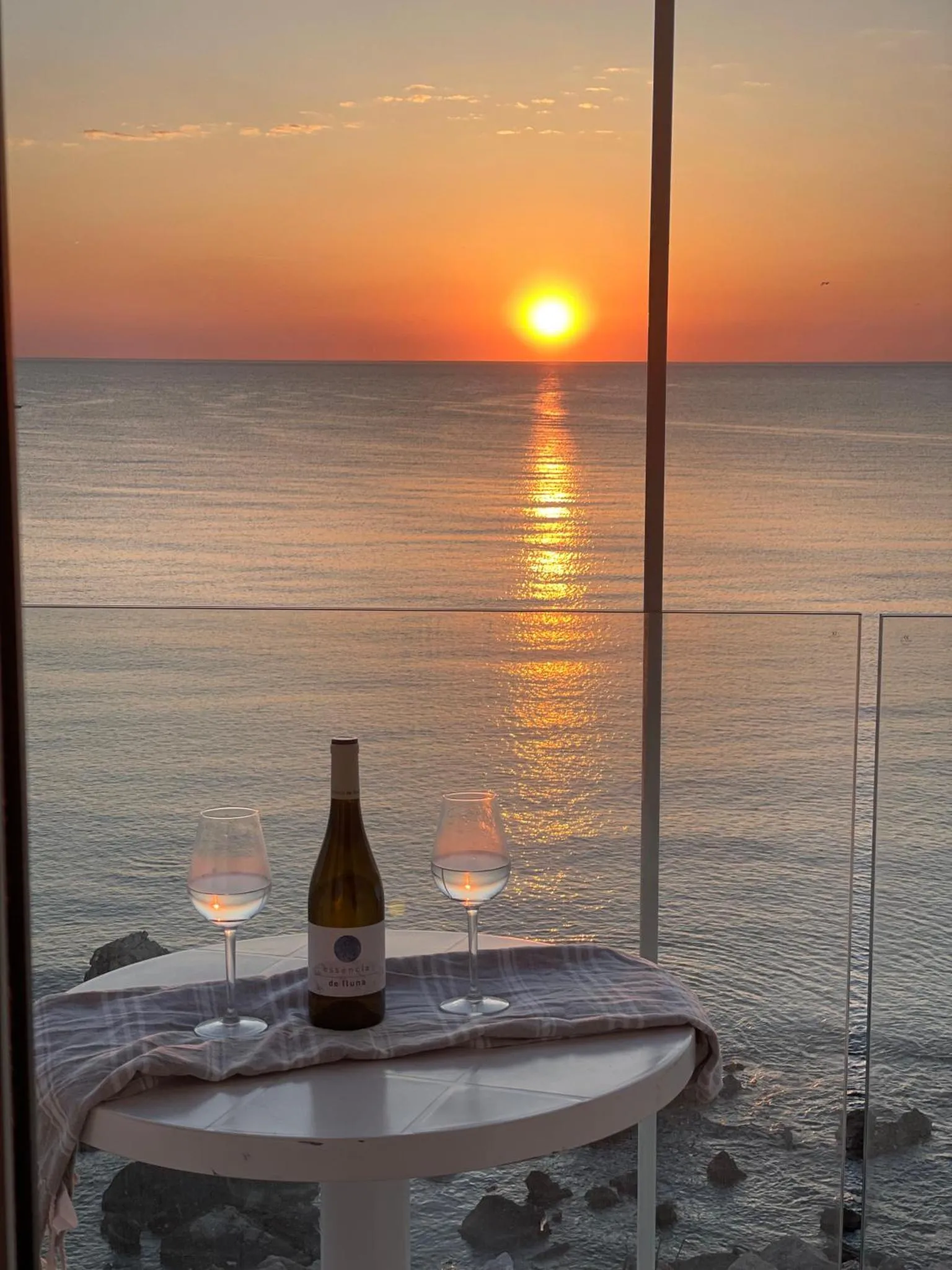 Une bouteille de vin et deux verres sur une table basse devant une vue sur la mer au coucher du soleil.