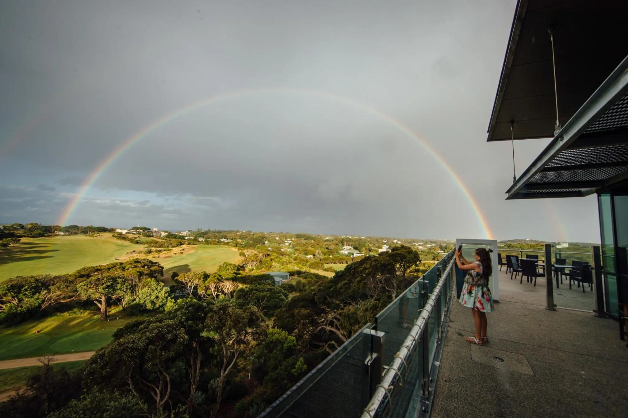 Une personne admire un arc-en-ciel depuis une terrasse avec vue sur un paysage verdoyant.