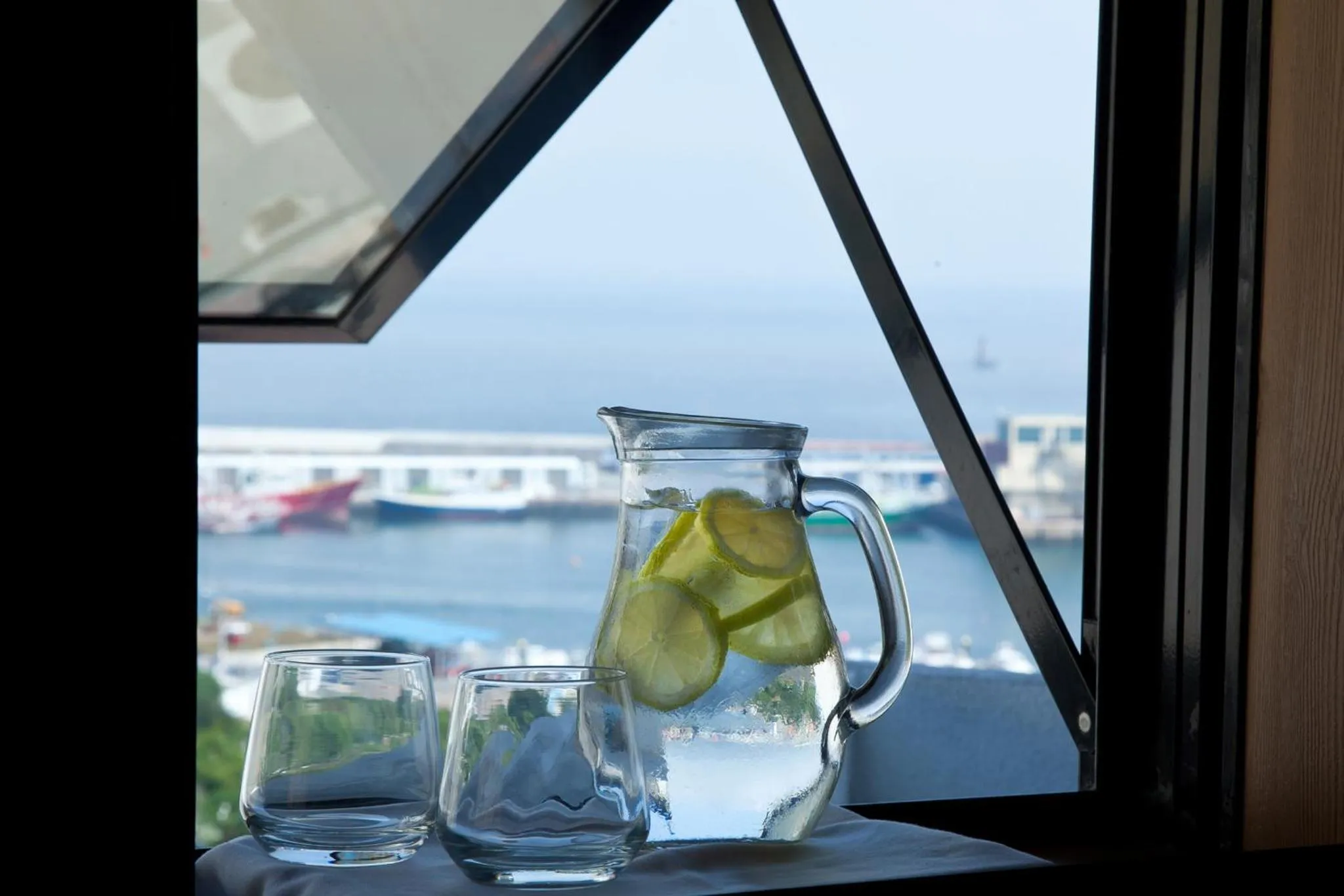 Une carafe d'eau fraîche et deux verres sont posés sur une surface sombre près d'une fenêtre ouverte sur la mer.