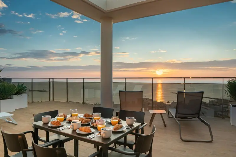 Une table de petit-déjeuner est dressée sur une terrasse avec vue sur la mer au coucher du soleil.