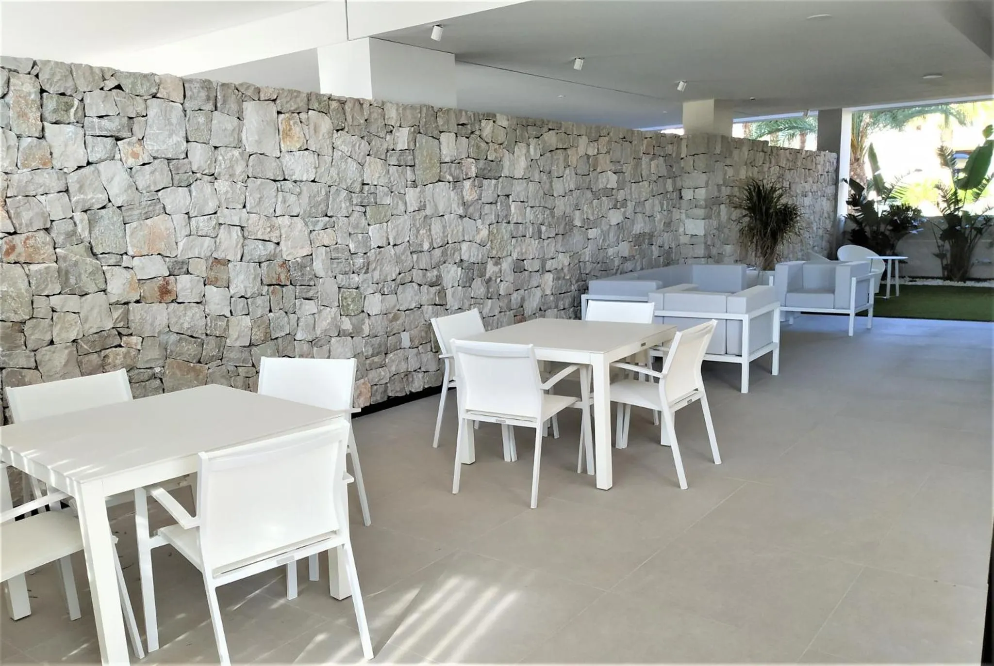 Espace repas extérieur avec tables et chaises blanches devant un mur en pierre.