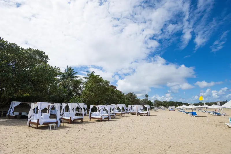 Transats blancs sous des rideaux sur une plage de sable fin, avec une végétation luxuriante en arrière-plan et un ciel bleu nuageux.
