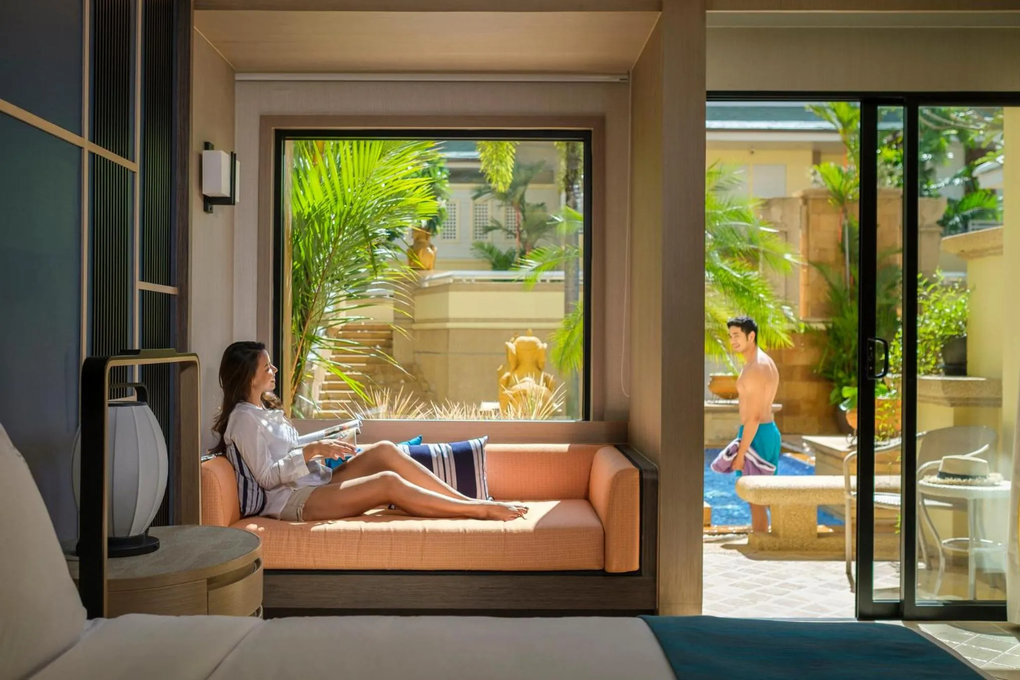 Une femme se détend sur un canapé près d'une fenêtre ouverte sur une terrasse et une piscine.