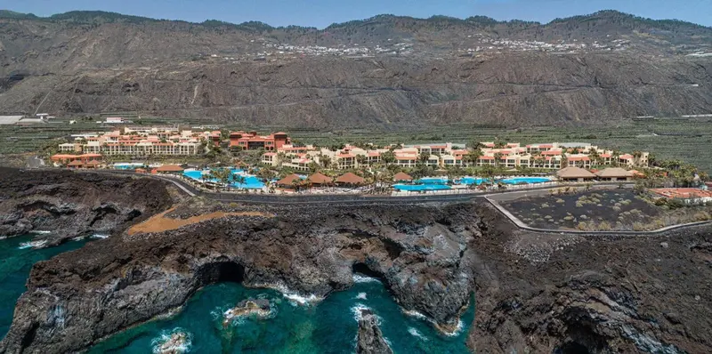 Vue aérienne d'un complexe hôtelier avec piscines et végétation luxuriante, surplombant une côte rocheuse et l'océan.
