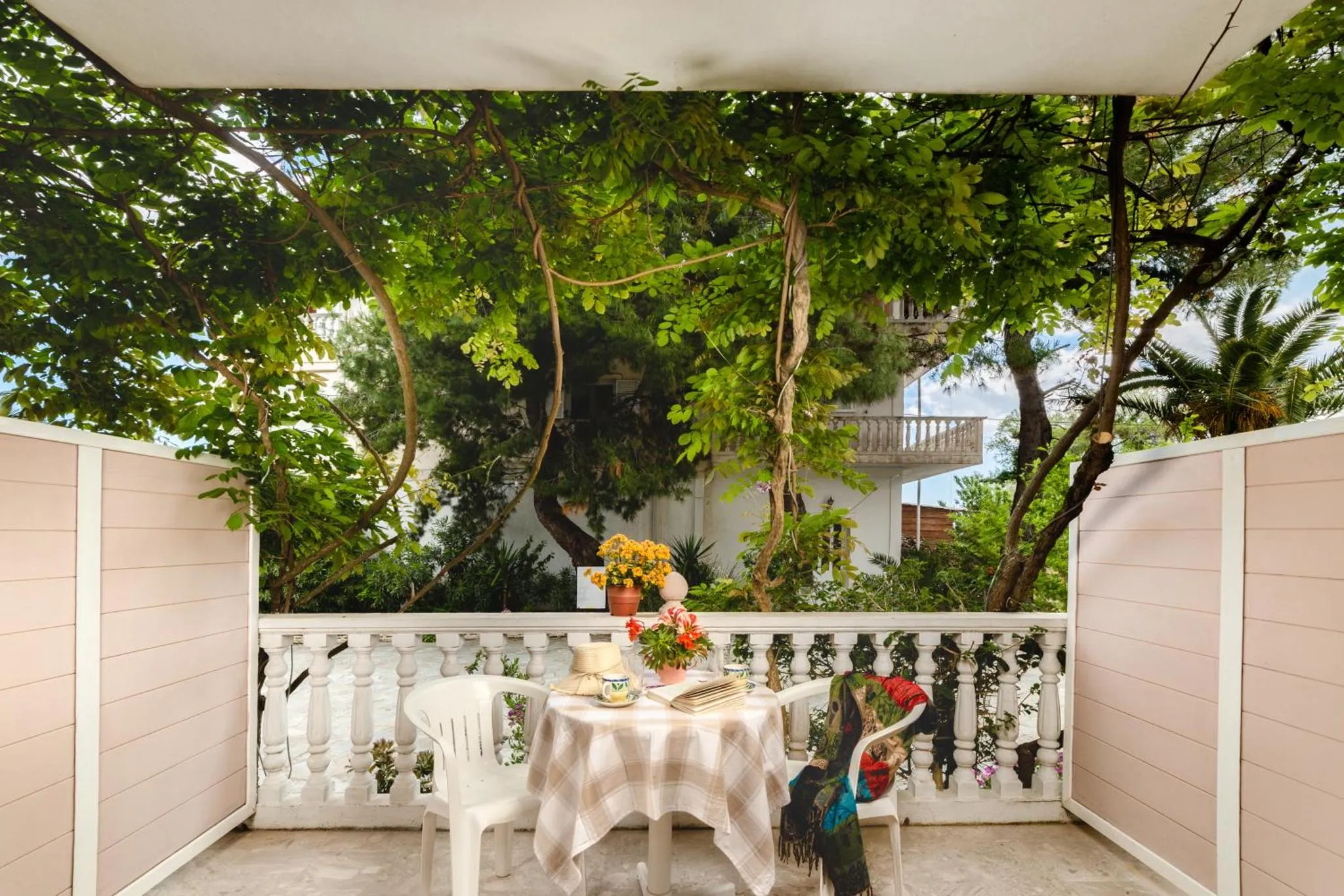 Une table et des chaises blanches sont disposées sur une terrasse ombragée par la végétation.
