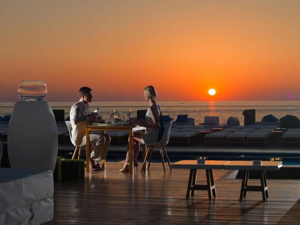 Un couple dîne à une table en terrasse face à la mer au coucher du soleil.