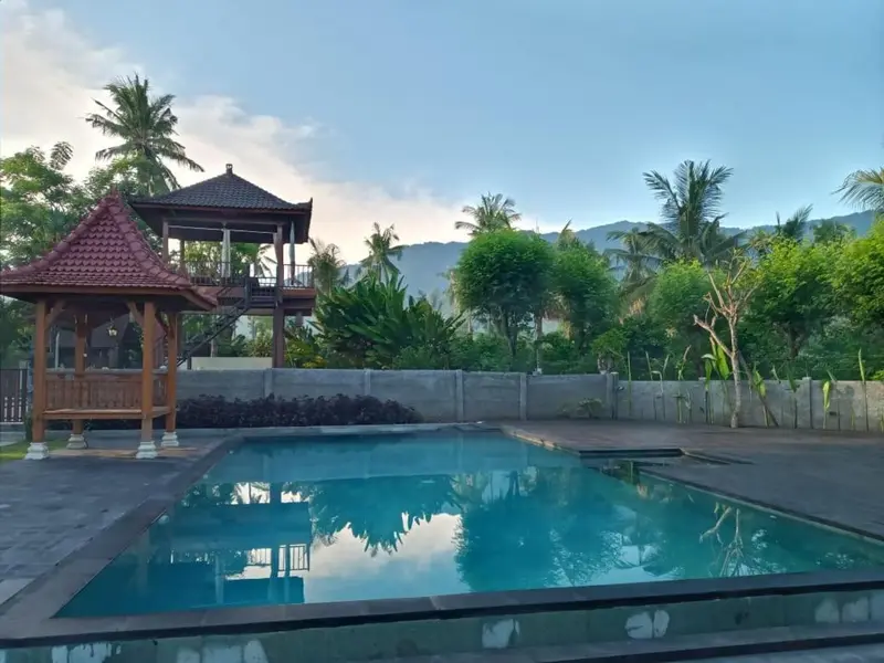 Une piscine rectangulaire turquoise est entourée d'une terrasse en bois et de végétation tropicale.