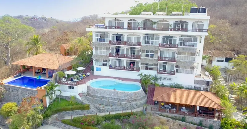 Bâtiment blanc à plusieurs étages avec des balcons surplombant des piscines et une végétation luxuriante.