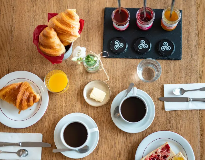 Un petit-déjeuner avec des viennoiseries, des confitures, du café et du jus d'orange est présenté sur une table en bois.