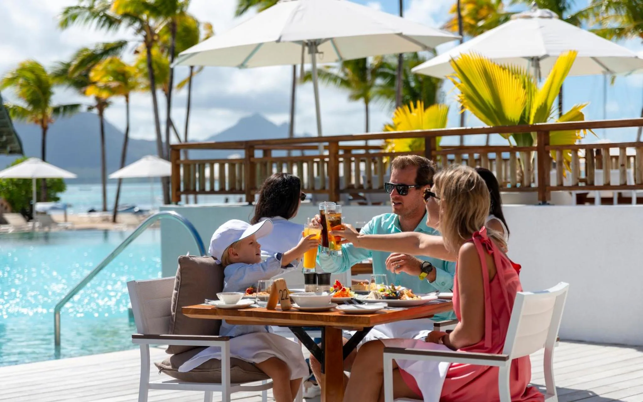 Une famille trinque avec des boissons fraîches sur une terrasse ensoleillée près d'une piscine.