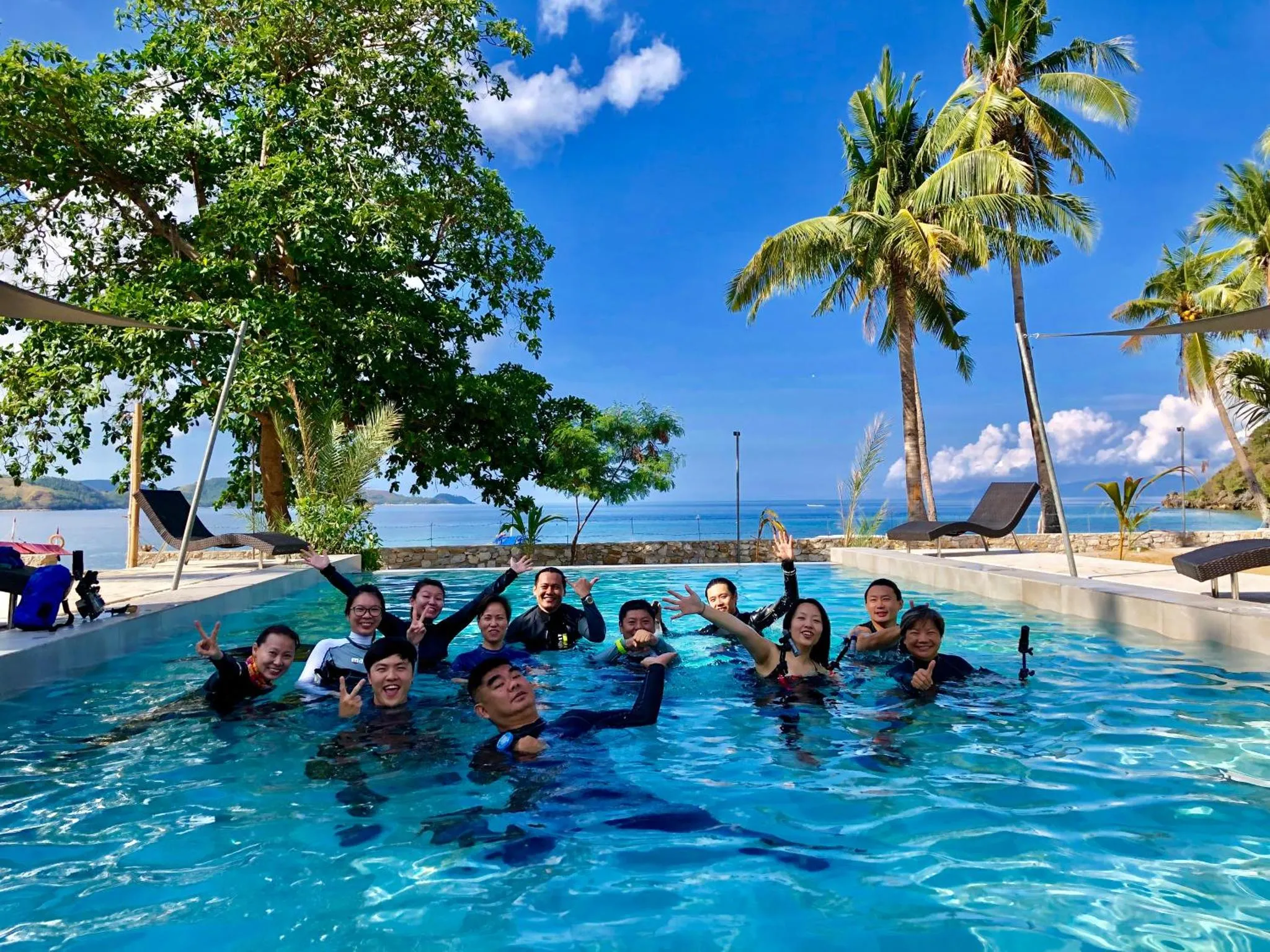 Groupe de personnes souriantes dans une piscine en bord de mer, sous les palmiers.
