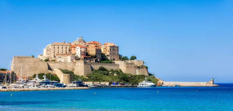 Vue d'ensemble d'une ville fortifiée surplombant un port de plaisance et la mer.
