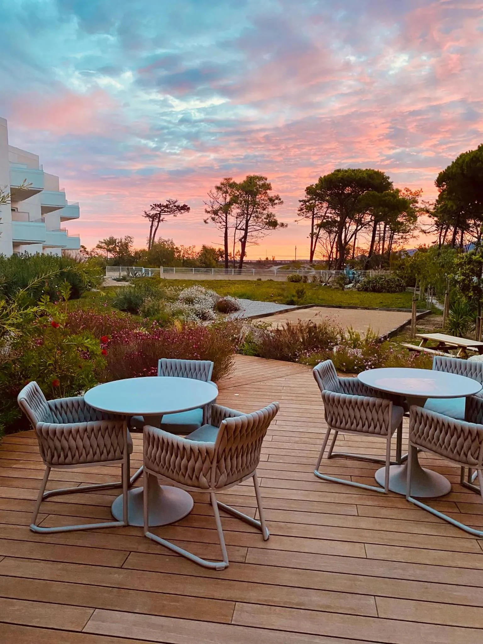 Tables et chaises sur une terrasse en bois avec végétation et coucher de soleil en arrière-plan.