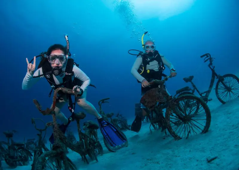 Deux plongeurs sourient devant une installation artistique de vélos rouillés au fond de l'océan.