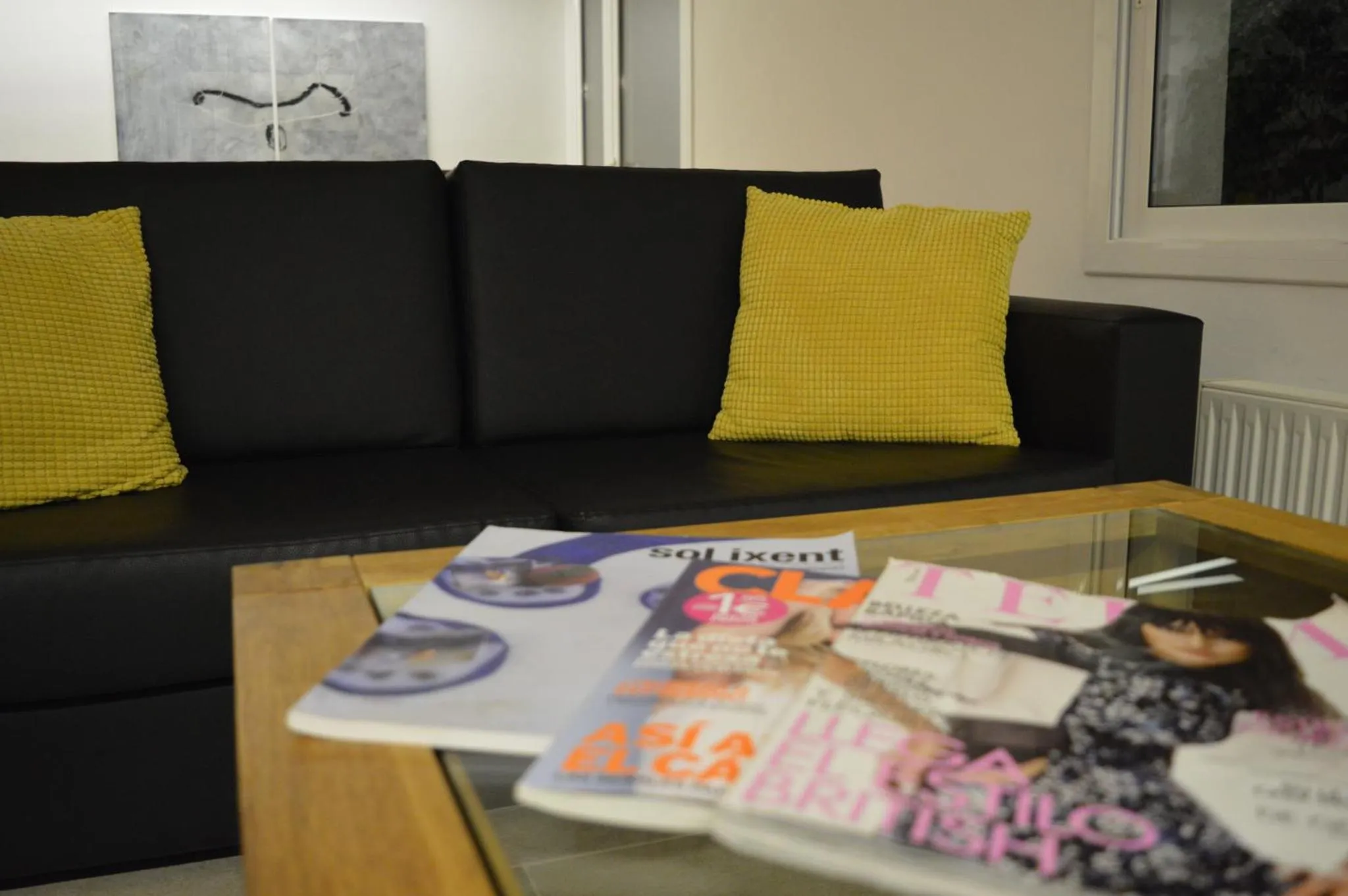 Un canapé noir avec des coussins jaunes devant une table basse en verre sur laquelle reposent des magazines.