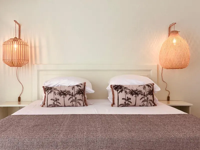 Un lit double avec des oreillers blancs et des coussins à motifs de palmiers, éclairé par deux lampes en rotin.