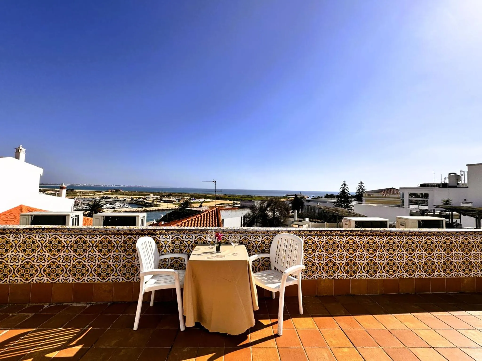 Table et chaises sur une terrasse carrelée avec vue sur la mer et le port.