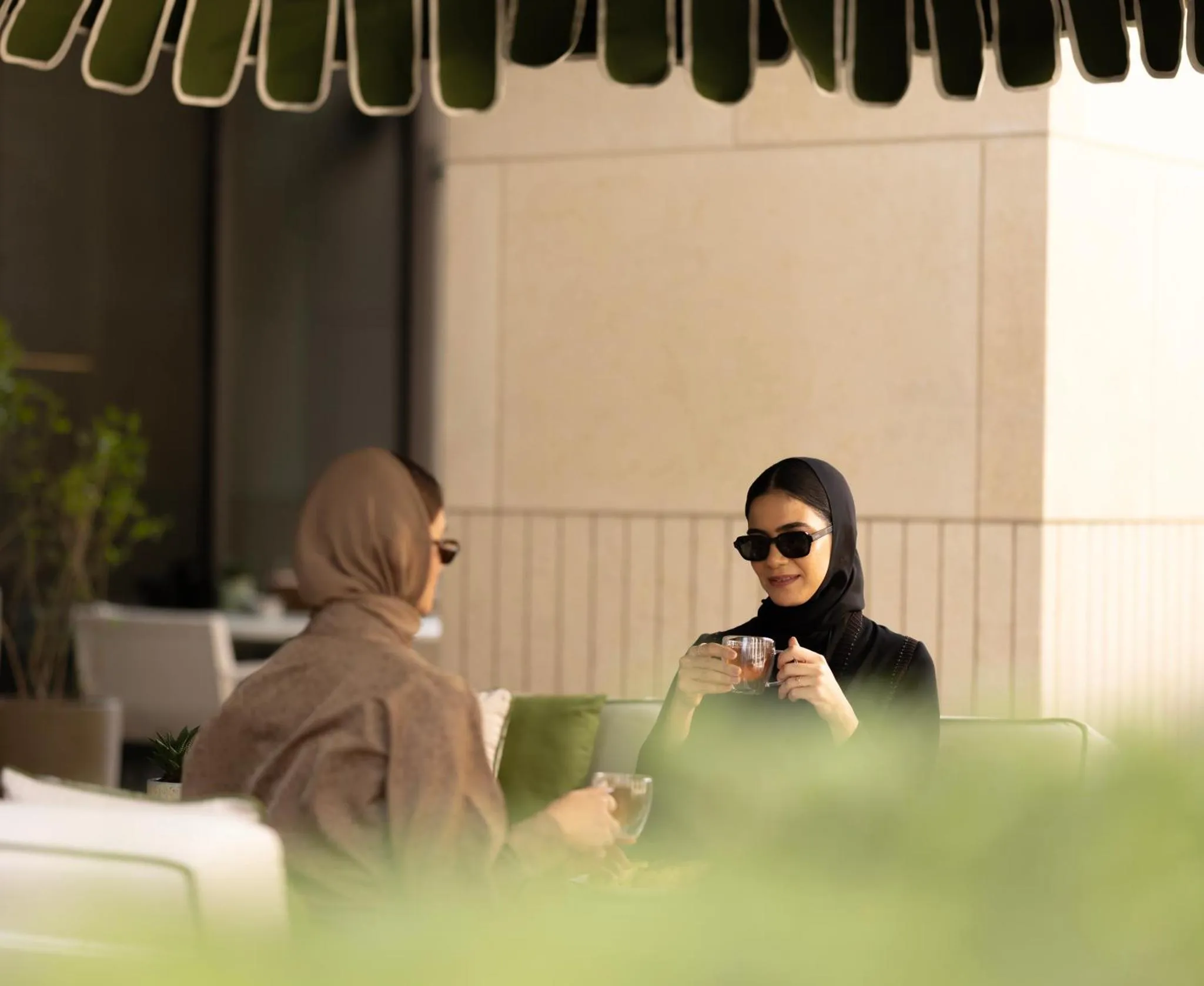 Deux femmes assises à une table boivent une boisson sur une terrasse ombragée.