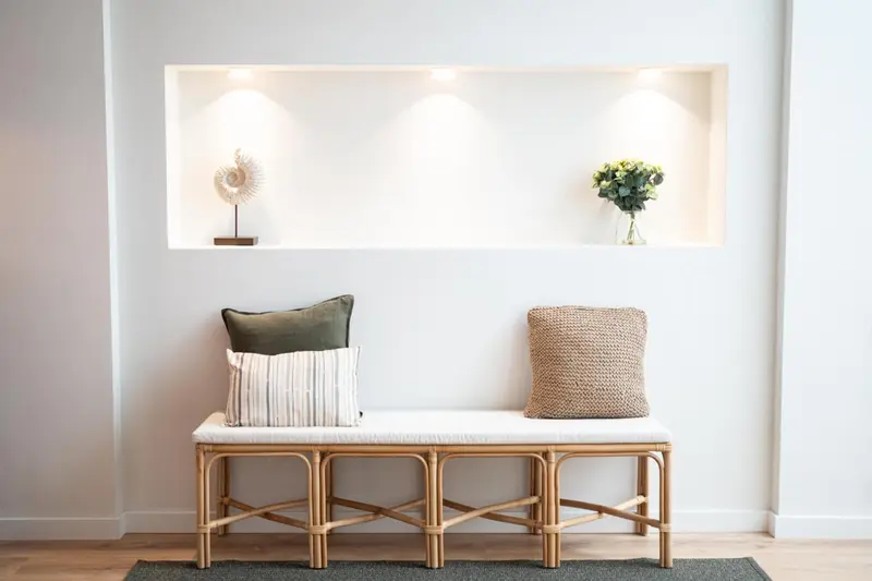 Banc en rotin avec coussins devant un mur blanc avec une niche décorative.