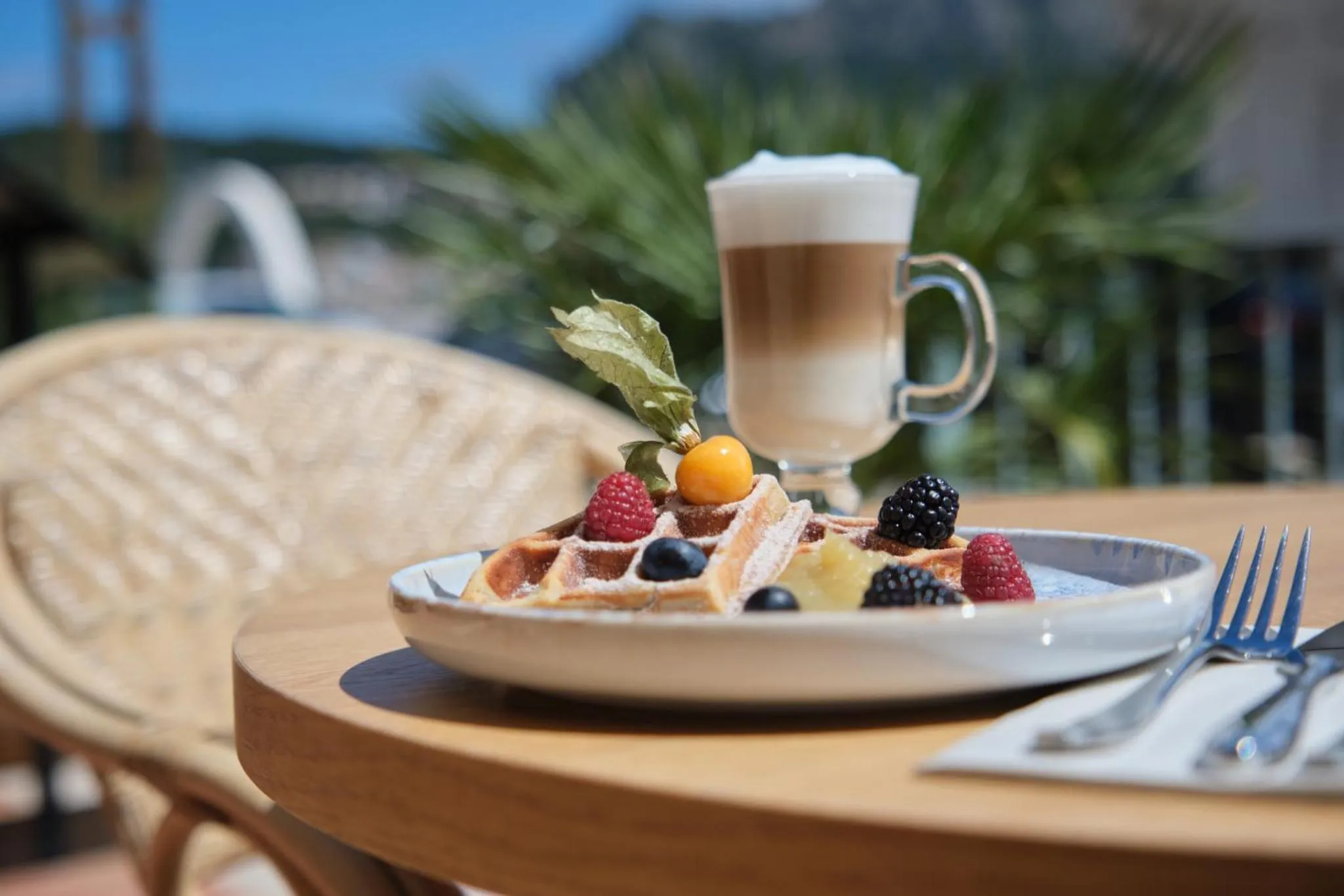 Un gaufre garni de fruits et accompagné d'un cappuccino est servi sur une table en terrasse.