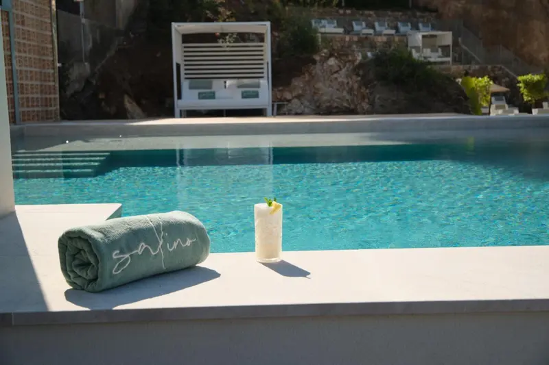Une serviette roulée et un cocktail sont posés sur le bord d'une piscine turquoise.