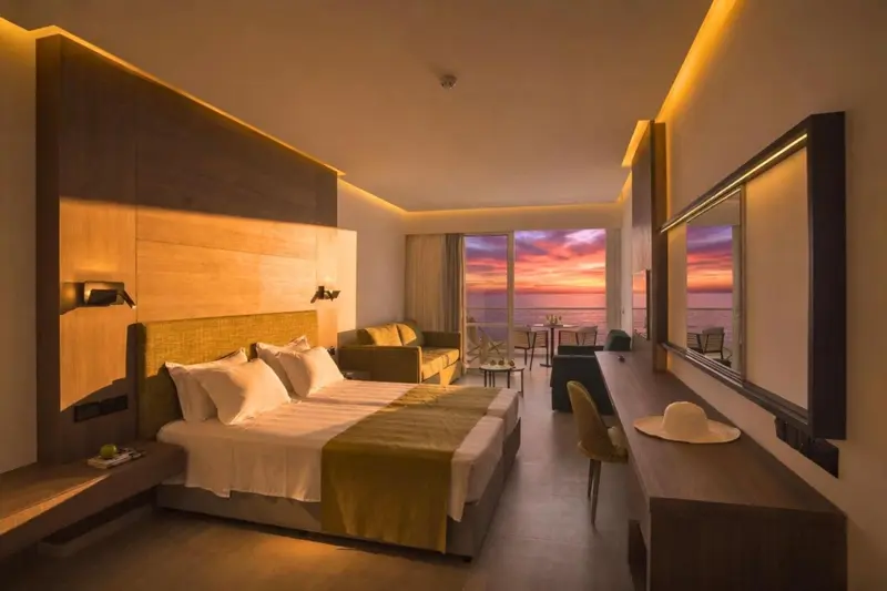 Chambre lumineuse avec un grand lit et une vue sur la mer au coucher du soleil.