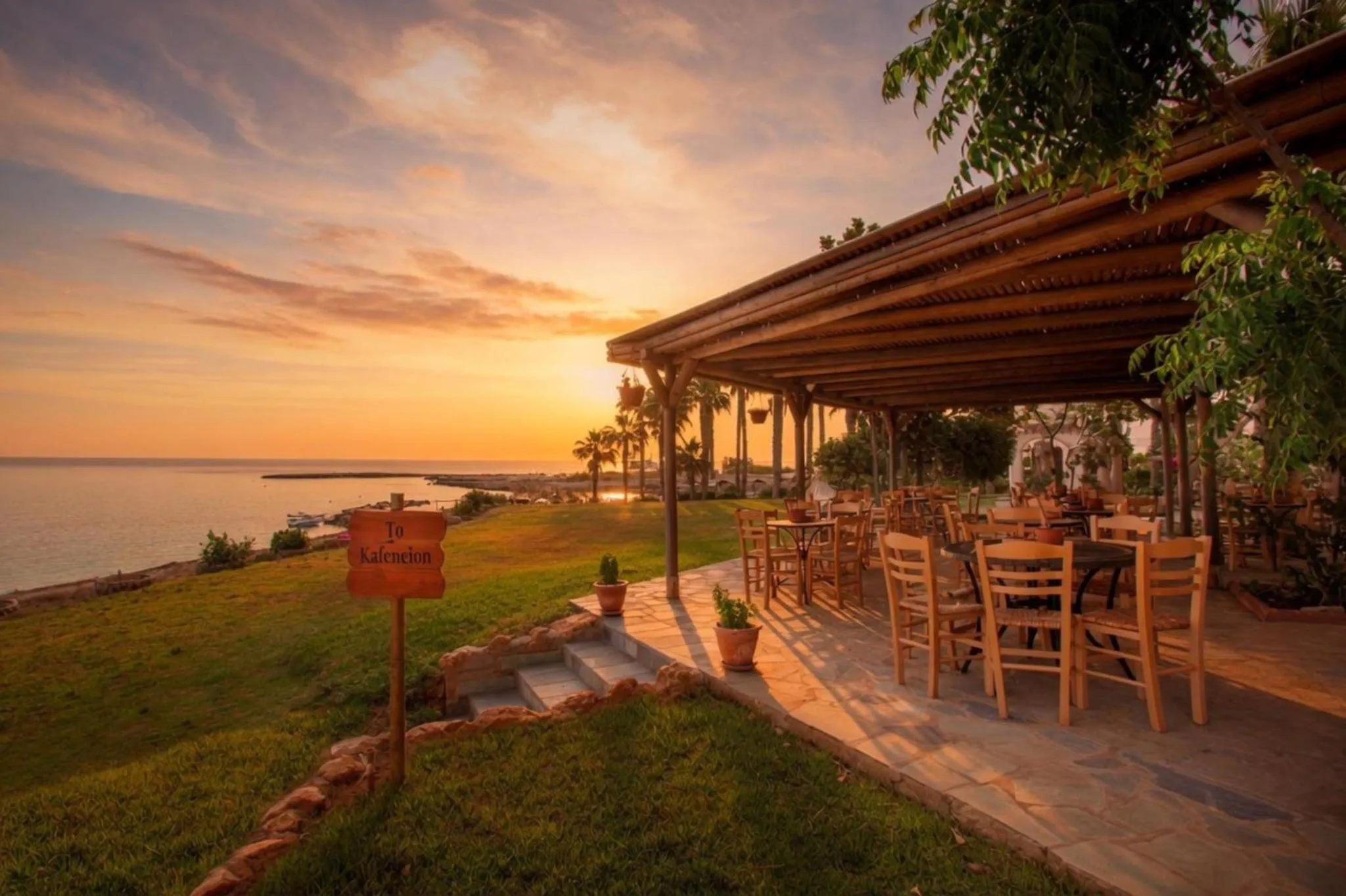 Terrasse ombragée avec tables et chaises donnant sur la mer au coucher du soleil.