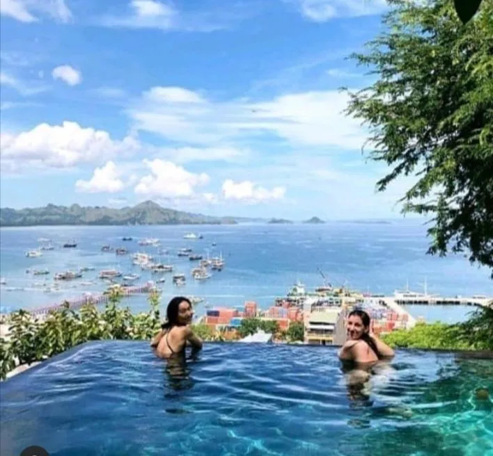 Deux femmes sourient dans une piscine à débordement avec vue sur la mer et des bateaux.